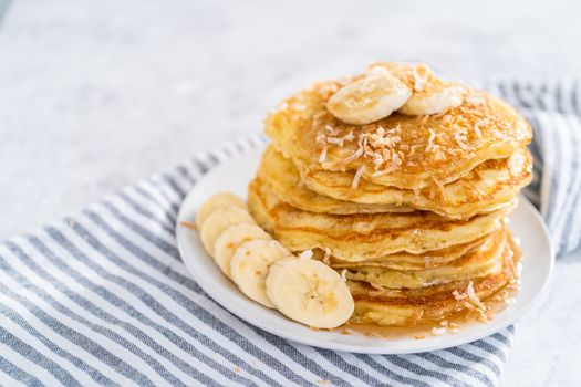 Stack of freshly baked coconut banana pancakes garnished with sliced bananas, toasted coconut, and maple syrup.