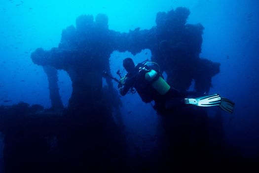 diver taking pictures of the remains of a sunken ship. The old remains of the ship are surrounded by lots of small fish and the blue and turbid water gives an aura of mystery