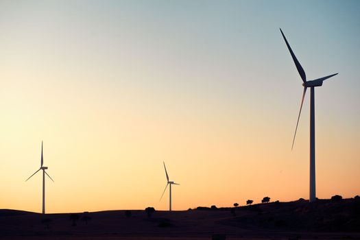 Silhouette of three windmills producing green energy in a wind farm at sunset. They are in a rural environment surrounded by crops and trees, the sky is clean and clear. Copy space
