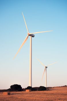 vertical photo of two windmills on a small hill producing clean energy in a wind farm at sunset. It is in a rural environment surrounded by crops and nature, the sky is clean and clear. Copy space