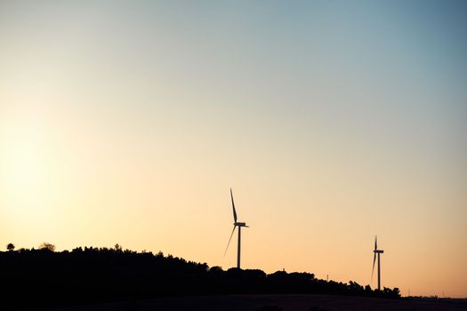 Silhouette of two windmills producing clean energy in a wind farm at sunset. They are in a rural environment surrounded by crops and trees, the sky is clean and clear. Copy space