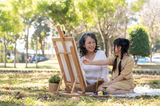 Happy and lovely Asian grandmother teaching her granddaughter to paint watercolors on canvas, having a great time together in the beautiful park.