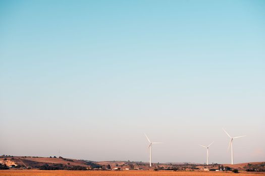 three windmills produce clean energy in a wind farm. They are in a rural environment surrounded by crops and small hills, the sky is clean and clear. Copy space