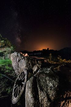 A wagon wheel is supported on the long wall of large stones of a farm. The dark night sky is full of stars and behind the mountains you can see the reflection of the lights of a nearby town