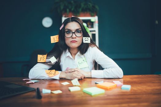 Crazy woman with reminder labels sitting at office desk. Many note paper stickers pasted on young Caucasian business woman. Multitasking concept. Deadline at work. Tinted image.