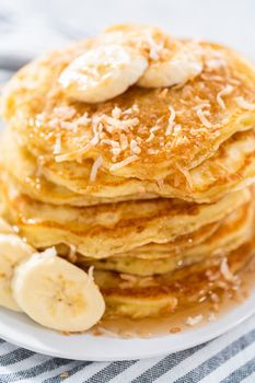 Stack of freshly baked coconut banana pancakes garnished with sliced bananas, toasted coconut, and maple syrup.