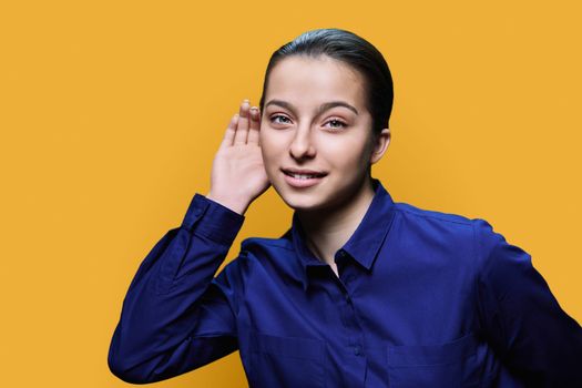 Young attractive teen female holding hand near ear, listening attentively, on yellow studio background. Secret conversation, news, gossip, eavesdropping concept