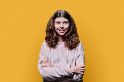 Portrait of smiling child schoolgirl on yellow studio background. Preteen girl looking at camera with crossed arms, wearing school clothes. School, kids, study education, childhood concept