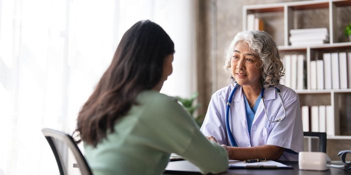 Woman senior doctor is Reading Medical History of Female Patient and Speaking with Her During Consultation in a Health Clinic. Physician in Lab Coat Sitting Behind a Laptop in Hospital Office...