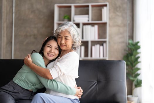 Loving adult daughter hugging older mother, standing behind couch at home, family enjoying tender moment together, young woman and mature mum or grandmother looking at each other, two generations..