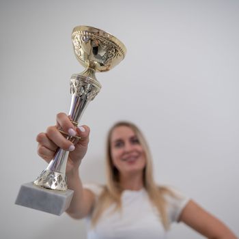 Blonde woman smiling and holding a golden trophy cup against a white wall