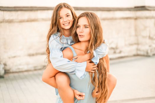 Mother of the daughter walks playing. Mother holds the girl on her back, holding her legs, and her daughter hugs her by the shoulders. Dressed in blue dresses