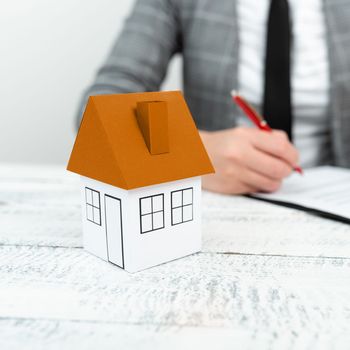 Businesswoman sitting and writing in notebook. Paper house on desk.