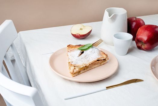 Autumn food. Homemade piece of apple pie with fresh red apples on white dining table, beige wall