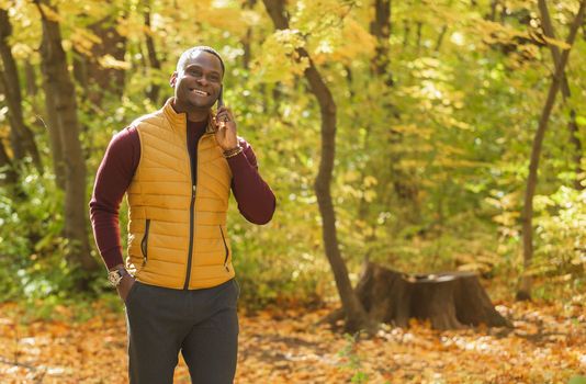 Portrait of smiling african american man talking on mobile phone outside in autumn park - communication and technologies concept