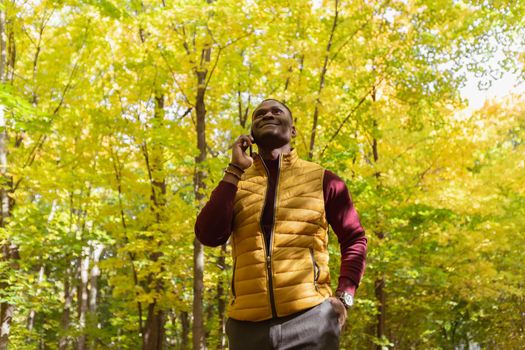 Portrait of smiling african american man talking on mobile phone outside in autumn park - communication and technologies concept