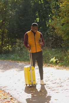 Cheerful african american young man in stylish clothes with suitcase walks in an autumn park on warm sunny day. Countryside travel concept