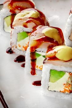 Closeup of sushi rolls with cream cheese, avocado and flying fish roe topped with butterfish fillet slices, mayonnaise and unagi sauce served on white plate. Japanese cuisine