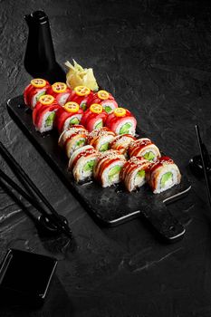 Set of Philadelphia rolls stuffed with cream cheese, avocado and cucumber topped with eel and tuna seasoned with unagi sauce garnished with slices of kumquat on black serving board with pickled ginger