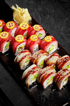 Two types of sushi rolls with cream cheese, avocado and cucumber topped with eel and tuna seasoned with unagi sauce garnished with slices of kumquat on black serving board with pickled ginger