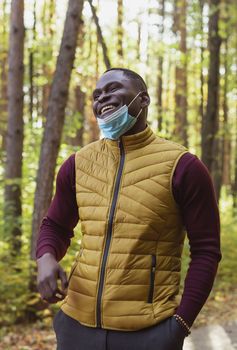 Handsome african american man wearing casual clothes and medical mask smiling happy walking in city park - end pandemic