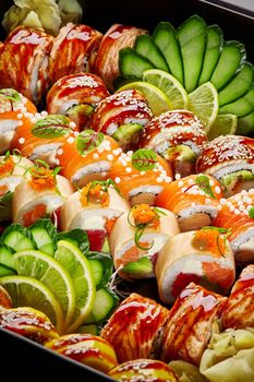 Closeup of various appetizing Japanese sushi rolls with pickled ginger, cucumber and lemon slices, greens packed in black cardboard box. Authentic cuisine. Delivery or takeaway food concept