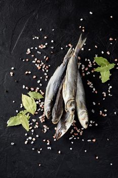 Bunch of dried mullet lying on black textured surface with scattered crystals of coarse Himalayan salt, peppercorns and dry bay leaves