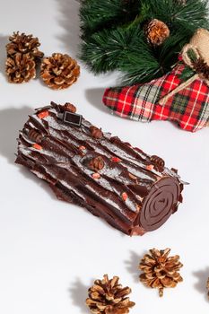 Delicious sweet cake roll in shape of log decorated with chocolate chips and candied berries on white background with green pine branch, fir cones and red checkered Christmas stocking. Festive treats