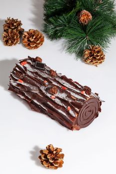 Log shaped sweet roll garnished with dark chocolate chips and candied berry crumbs on white background with Christmas tree branch and fir cones frosted with gold. Festive dessert concept