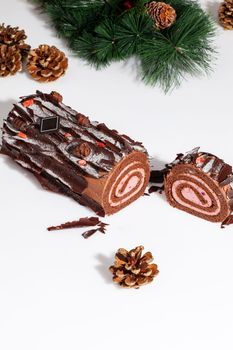 Sliced sweet chocolate sponge cake roll in shape of log with tender berry mousse filling covered with chocolate flakes on white surface with green Christmas tree branch and gilded cones