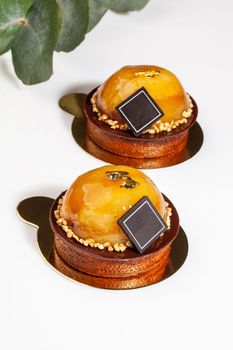 Two fresh sweet pastries on shortbread tartlet topped with chocolate and caramelized apples decorated with nut crumbs on golden serving cardboard on white background with green flower branch