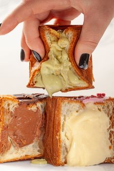 Closeup of female hand squeezing half of fresh soft croissant cube with delicate creamy pistachio custard filling, two more halves with vanilla and chocolate creams lying on table. Craving for sweets