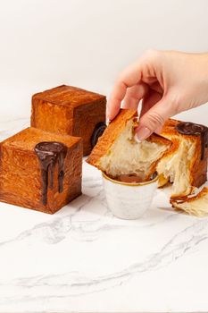 Female hands dipping sliced fresh fluffy cube shaped croissant into chocolate custard cream, two more whole cubic croissants with browned crust lying on table. Tempting French style puff pastries