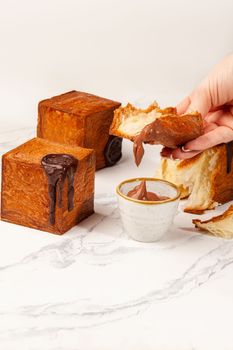 Woman holding slice of fresh flaky croissant with tender sweet hazelnut cream, two more whole cubic croissants decorated with chocolate seal on browned crust lying on table. French style pastries