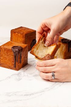 Woman hands dipping slice of soft flaky cube shaped croissant into small bowl with creamy chocolate custard on background of whole cubic croissants decorated with chocolate seal. Sweet tooth concept