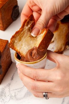 Closeup of female hands dipping piece of fresh fluffy cubic croissant with browned crispy crust into delicate chocolate cream in small bowl. Popular sweets concept. French style viennoiserie pastries