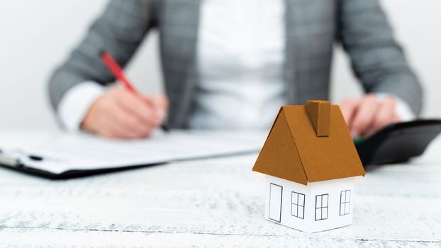 Businesswoman sitting and writing in notebook. Paper house' on desk.