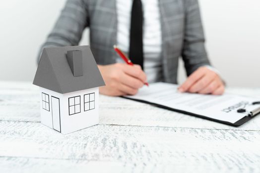 Businesswoman sitting and writing in notebook. Paper house on desk.