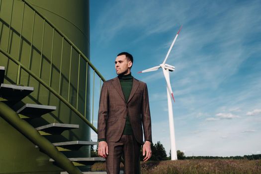 A man in a business suit with a green Golf shirt stands next to a windmill against the background of the field and the blue sky.Businessman near the windmills.Modern concept of the future