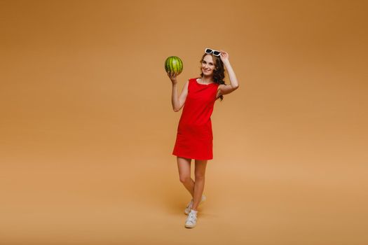 A beautiful girl in glasses and a red dress holds a watermelon in her hands.