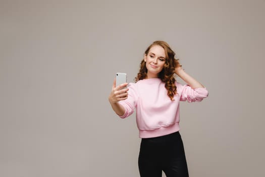 A smiling happy girl in a pink blouse takes a selfie on a smartphone on a gray background