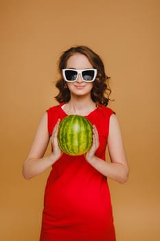 A beautiful girl in glasses and a red dress holds a watermelon in her hands.