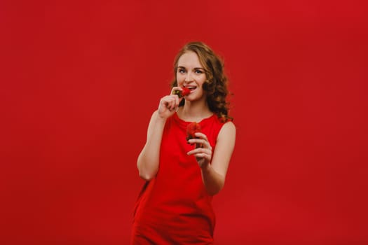 A beautiful girl in a red dress on a red background holds a strawberry in her hands and smiles.