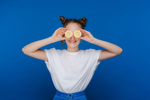 a young beautiful girl standing on a blue background holds lemons in her hands and covers her eyes with them.