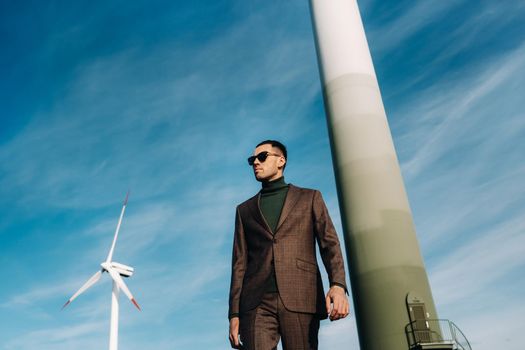 A man in a business suit with a green Golf shirt stands next to a windmill against the background of the field and the blue sky.Businessman near the windmills.Modern concept of the future