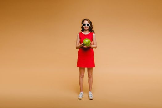 A beautiful girl in glasses and a red dress holds a watermelon in her hands.