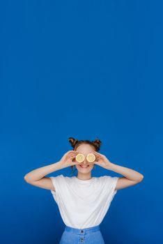 a young beautiful girl standing on a blue background holds lemons in her hands and covers her eyes with them.