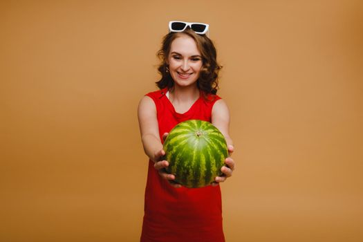 A beautiful girl in glasses and a red dress holds a watermelon in her hands.