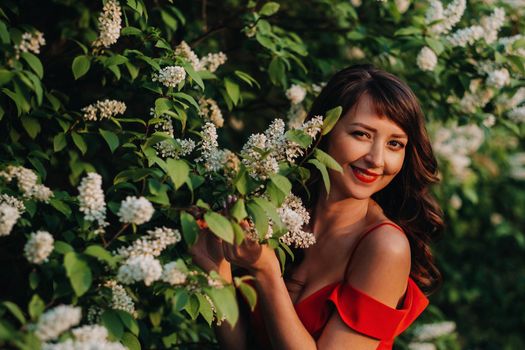 a girl in a red dress with red lips stands next to a large white flowering tree At sunset.