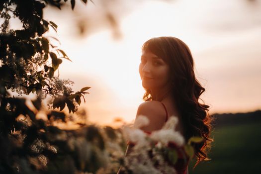 a girl in a red dress with red lips stands next to a large white flowering tree At sunset.